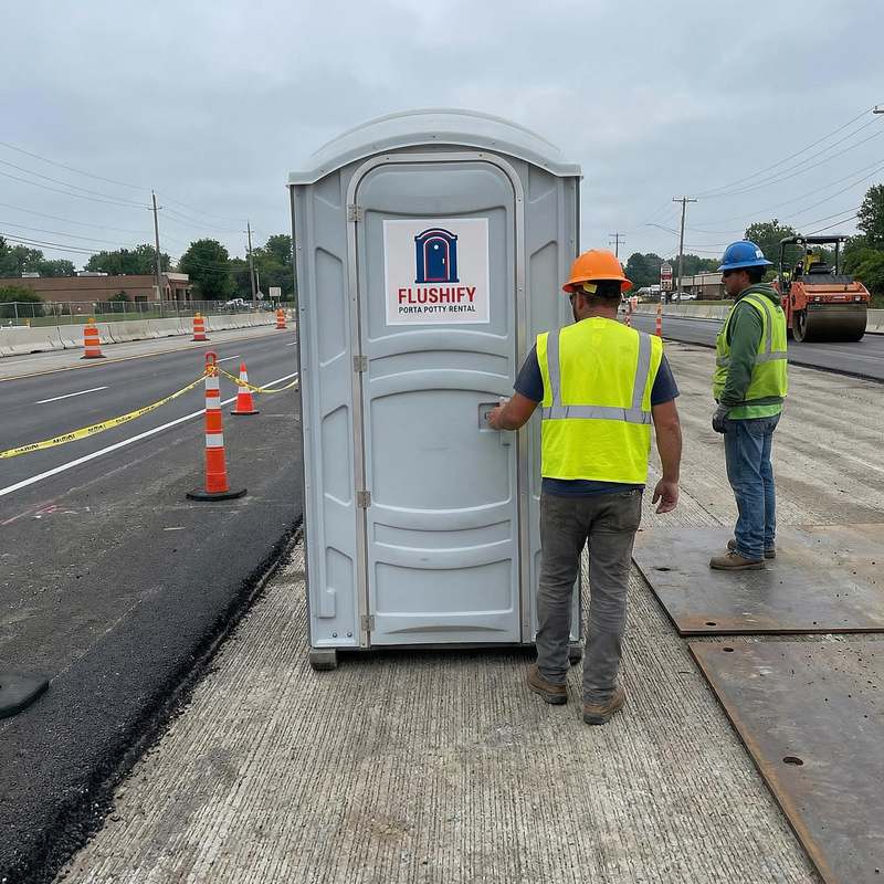 Grey portable toilet beside a road crew in safety vests